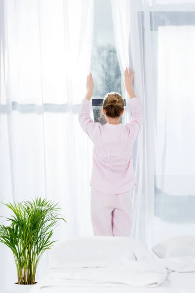 Vista trasera de la mujer en pijama abriendo cortinas en la mañana - foto de stock