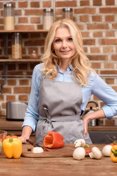 Lächelnde Frau steht am Tisch in der Küche und blickt in die Kamera — Stockfoto