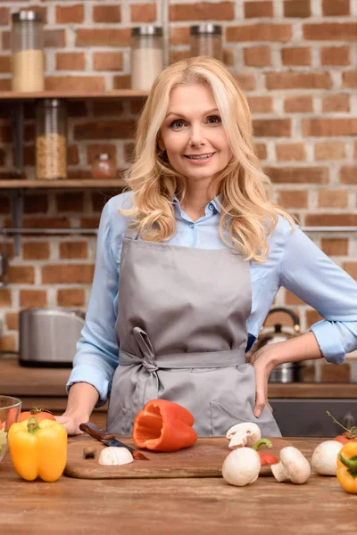 Sonriente mujer de pie cerca de la mesa en la cocina y mirando a la cámara - foto de stock