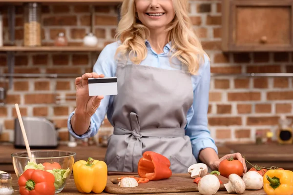 Zugeschnittenes Bild einer Frau, die Kreditkarte zeigt und in der Küche neben einem Tisch mit Gemüse steht — Stockfoto