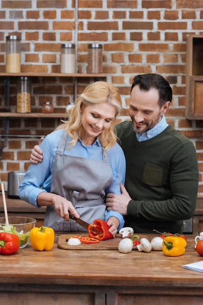 Чоловік обіймає дружину, коли вона ріже овочі на кухні — стокове фото