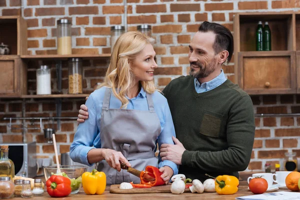 Муж обнимает жену, пока она режет овощи на кухне — стоковое фото