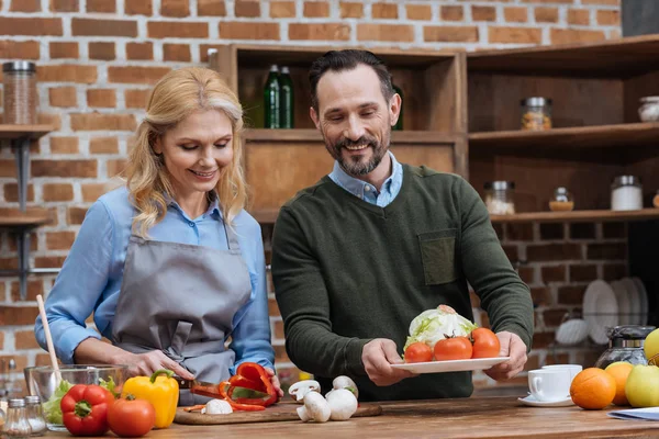 Esposa cortar verduras y marido poner plato en la mesa - foto de stock
