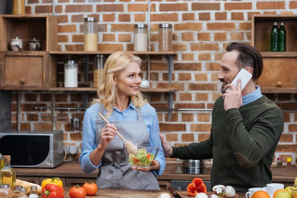 Sorrindo esposa misturando salada na cozinha e marido falando por smartphone — Fotografia de Stock