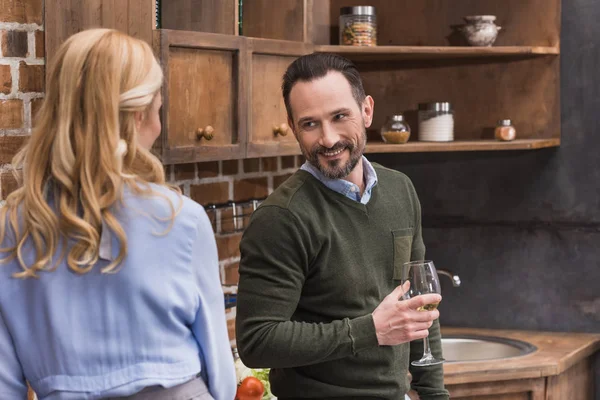 Улыбающийся муж с бокалом вина разговаривает с женой на кухне — стоковое фото