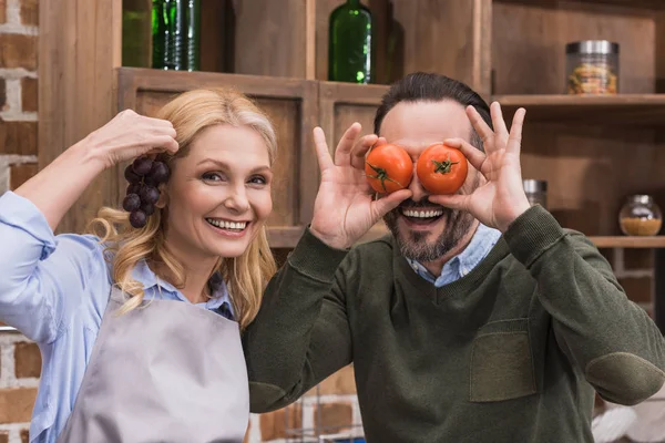 Esposa e marido se divertindo e sorrindo com pedaços de vegetais e frutas — Fotografia de Stock