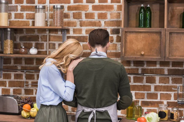 Задний вид жены обнимающей мужа, пока он готовит на кухне — стоковое фото