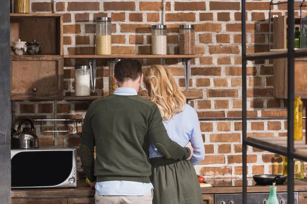 Задний вид мужа обнимающего жену на кухне — стоковое фото