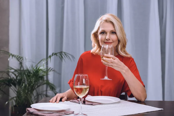 Atractiva mujer en vestido rojo beber vino en la mesa - foto de stock