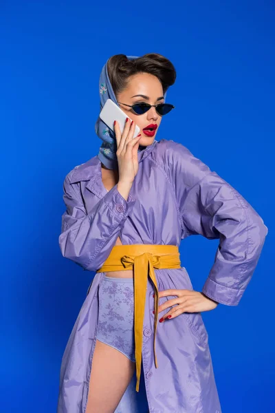 Retrato de mujer de moda en ropa de estilo retro hablando en teléfono inteligente aislado en azul - foto de stock