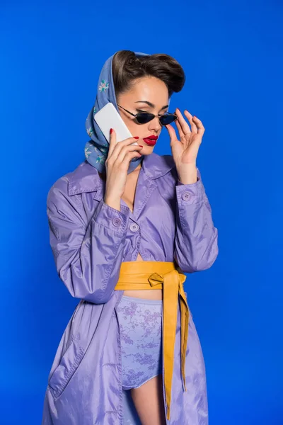Retrato de mujer de moda en ropa de estilo retro hablando en teléfono inteligente aislado en azul - foto de stock