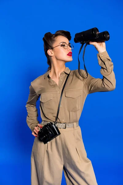 Portrait of stylish young woman with binoculars and photo camera isolated on blue — Stock Photo