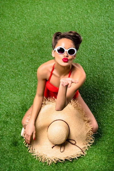 Overhead view of stylish young woman in swimming suit with straw hat blowing kiss on green grass — Stock Photo