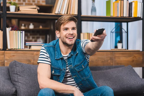 Sorrindo jovem usando controlador remoto enquanto sentado no sofá em casa — Fotografia de Stock