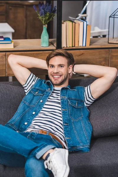 Bello giovane seduto sul divano e sorridente alla macchina fotografica — Foto stock