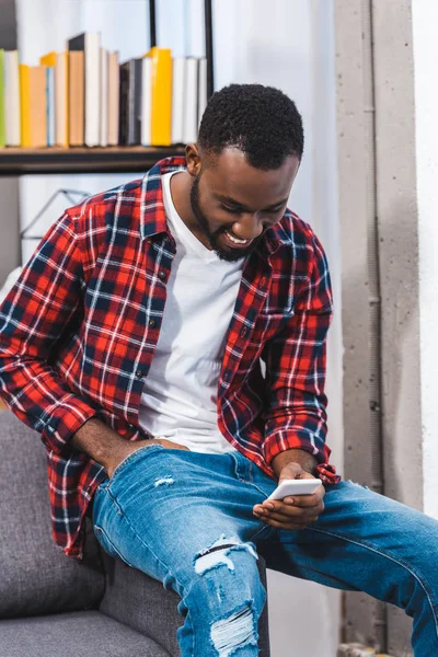 Sorridente giovane uomo africano americano utilizzando smartphone a casa — Foto stock