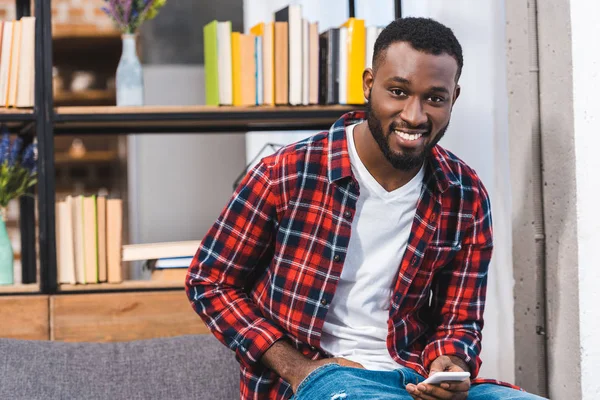 Guapo joven afroamericano hombre sosteniendo teléfono inteligente y sonriendo a la cámara - foto de stock