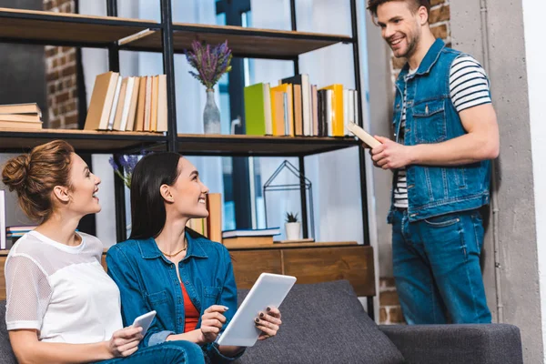 Souriant jeune homme tenant livre et regardant les filles en utilisant des appareils numériques — Photo de stock