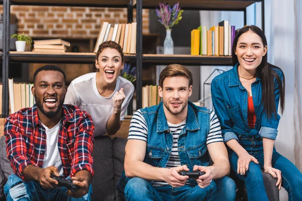 Cheerful young multiethnic friends playing with joysticks together at home — Stock Photo