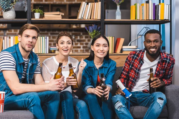 Счастливые молодые многонациональные друзья пьют пиво, сидя вместе на диване — стоковое фото