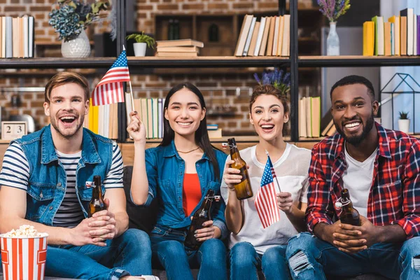 Felices jóvenes amigos multiétnicos sosteniendo banderas americanas y bebiendo cerveza en casa - foto de stock