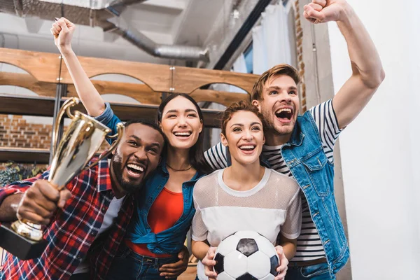 Aufgeregte junge multiethnische Freunde mit Fußball und Trophäenjubel zu Hause — Stockfoto