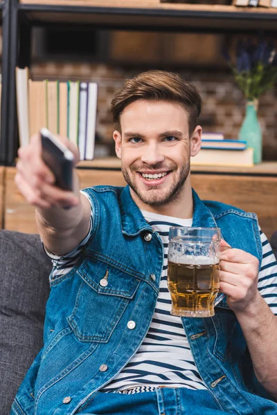 Sonriente joven sosteniendo un vaso de cerveza y un mando a distancia - foto de stock