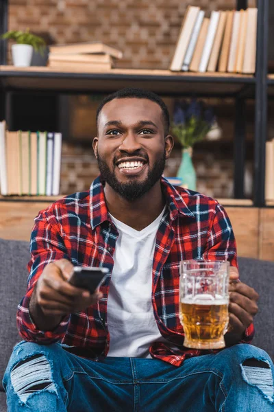Alegre joven afroamericano hombre sosteniendo vaso de cerveza y controlador remoto en casa - foto de stock