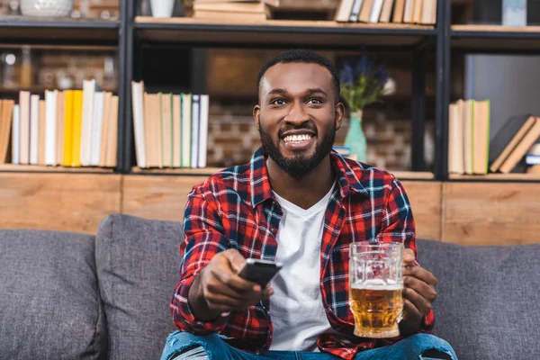Feliz jovem afro-americano segurando copo de cerveja e controle remoto enquanto sentado no sofá em casa — Fotografia de Stock