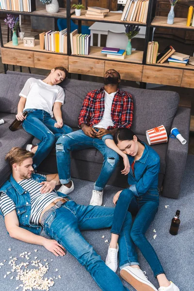 Vista de ángulo alto de jóvenes amigos multiétnicos que duermen en una habitación desordenada con palomitas de maíz y botellas de cerveza - foto de stock