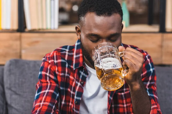 Jovem afro-americano bebendo cerveja enquanto sentado no sofá em casa — Fotografia de Stock