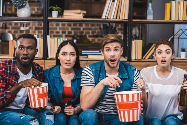 Chocado jovens amigos multiétnicos comer pipocas e beber cerveja enquanto assiste tv — Fotografia de Stock