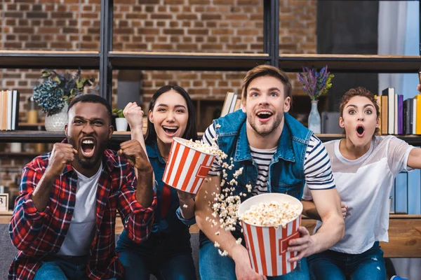 Emocional jóvenes amigos multiétnicos comiendo palomitas de maíz y viendo la televisión - foto de stock