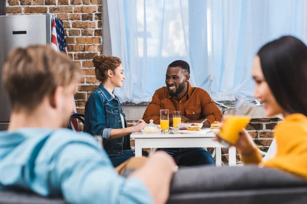 Junges lächelndes multiethnisches Paar sitzt am Tisch und Freunde trinken Saft im Vordergrund — Stockfoto
