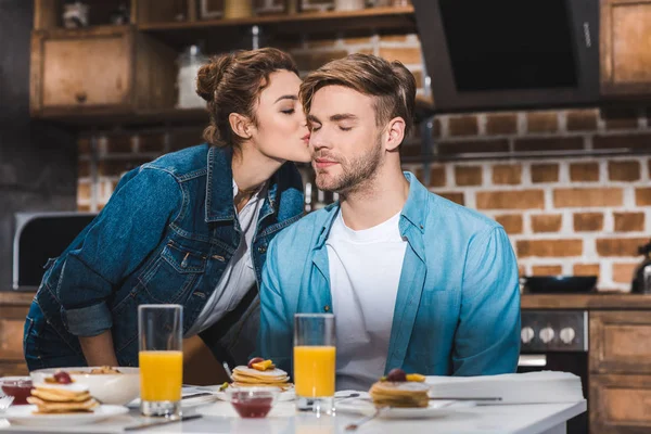 Menina beijando namorado sentado à mesa com suco em óculos e panquecas — Fotografia de Stock