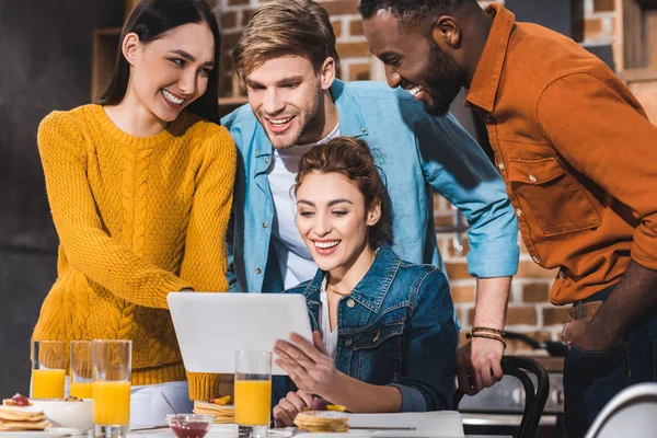 Smiling young multiethnic friends using digital tablet at table with juice and pancakes — Stock Photo