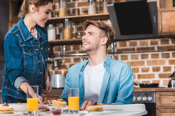 Feliz jovem casal sorrindo uns aos outros enquanto tomando café da manhã juntos em casa — Fotografia de Stock