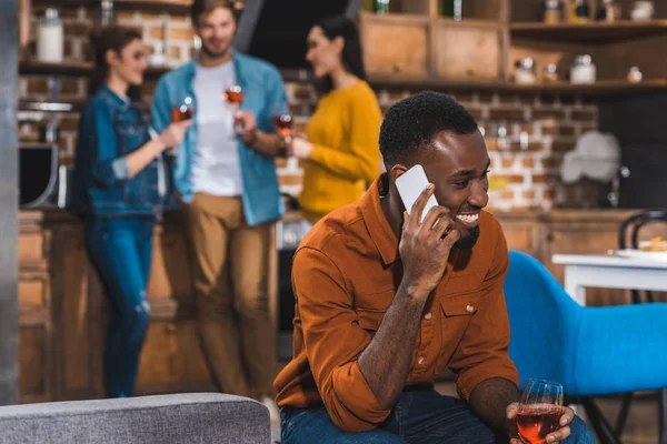 Feliz joven afroamericano hombre hablando por teléfono inteligente mientras amigos beber vino detrás - foto de stock