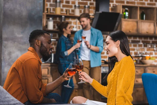 Vista lateral do jovem casal multiétnico beber vinho enquanto amigos de pé atrás — Fotografia de Stock