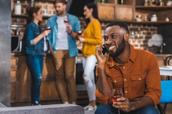 Sonriente afroamericano hombre hablando por teléfono inteligente mientras amigos bebiendo vino detrás - foto de stock