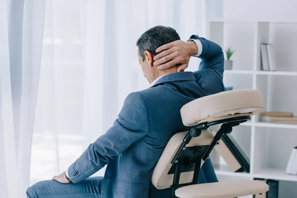 Adult businessman with painful backache sitting in massage chair at office — Stock Photo