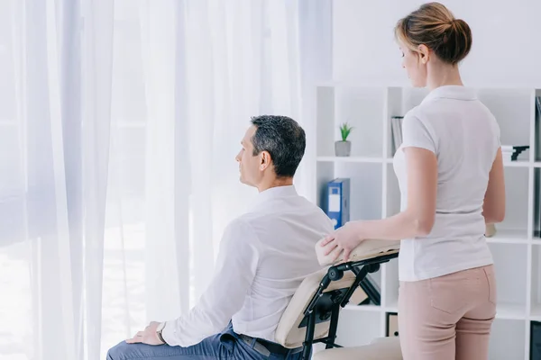 Geschäftsmann sitzt im Büro auf Massagesessel mit Masseurin hinter ihm — Stockfoto