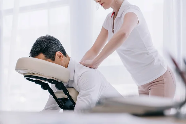 Masajista haciendo masaje de espalda sentado para hombre de negocios en la oficina - foto de stock