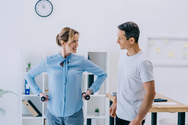 Feliz mujer de negocios adulta y su entrenador personal haciendo ejercicio con pesas en la oficina - foto de stock