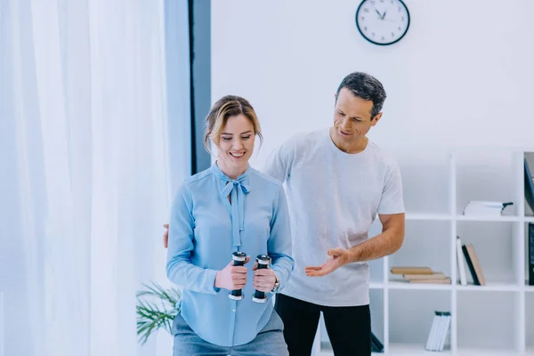 Feliz mujer de negocios haciendo ejercicio con pesas con entrenador en la oficina - foto de stock