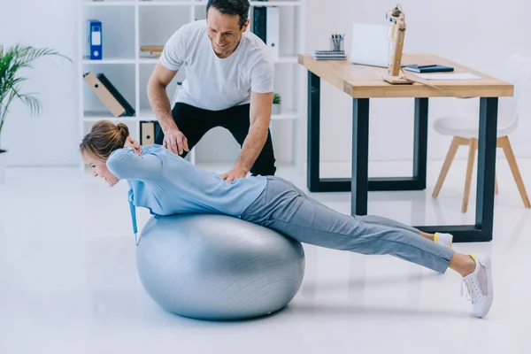 Side view of businesswoman doing hyperextension exercise on fit ball with trainer at office — Stock Photo