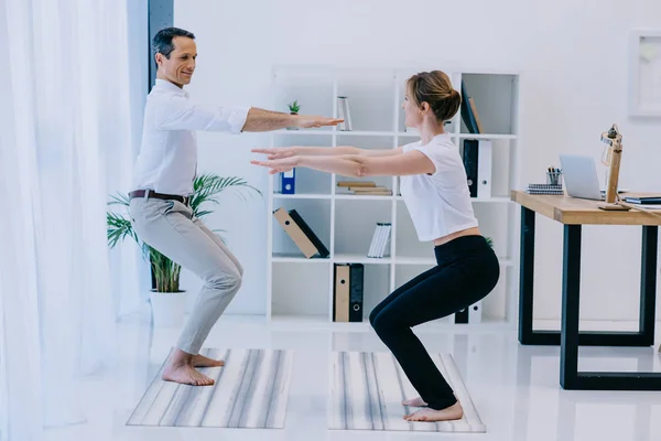 Vista lateral del hombre de negocios con su entrenador practicando yoga en pose incómoda en la oficina - foto de stock