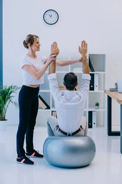 Businessman doing exercise while sitting on fit ball with female personal trainer — Stock Photo