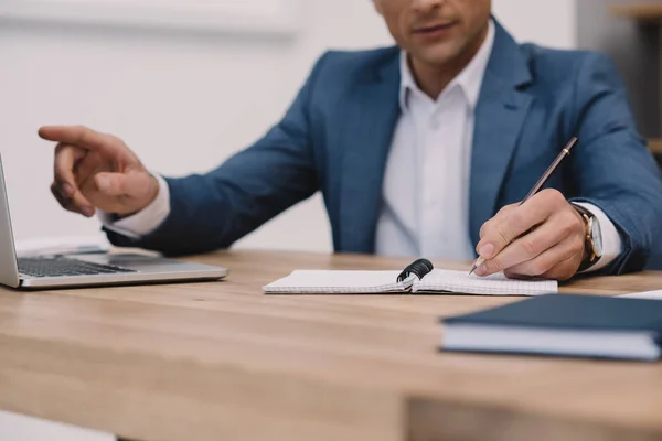 Cropped shot of businessman making notes while using laptop at workplace — Stock Photo