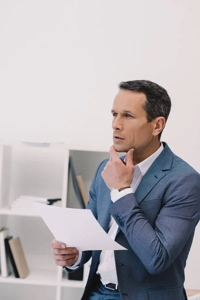Homme d'affaires réfléchi avec du papier d'affaires dans la main regardant loin au bureau — Photo de stock